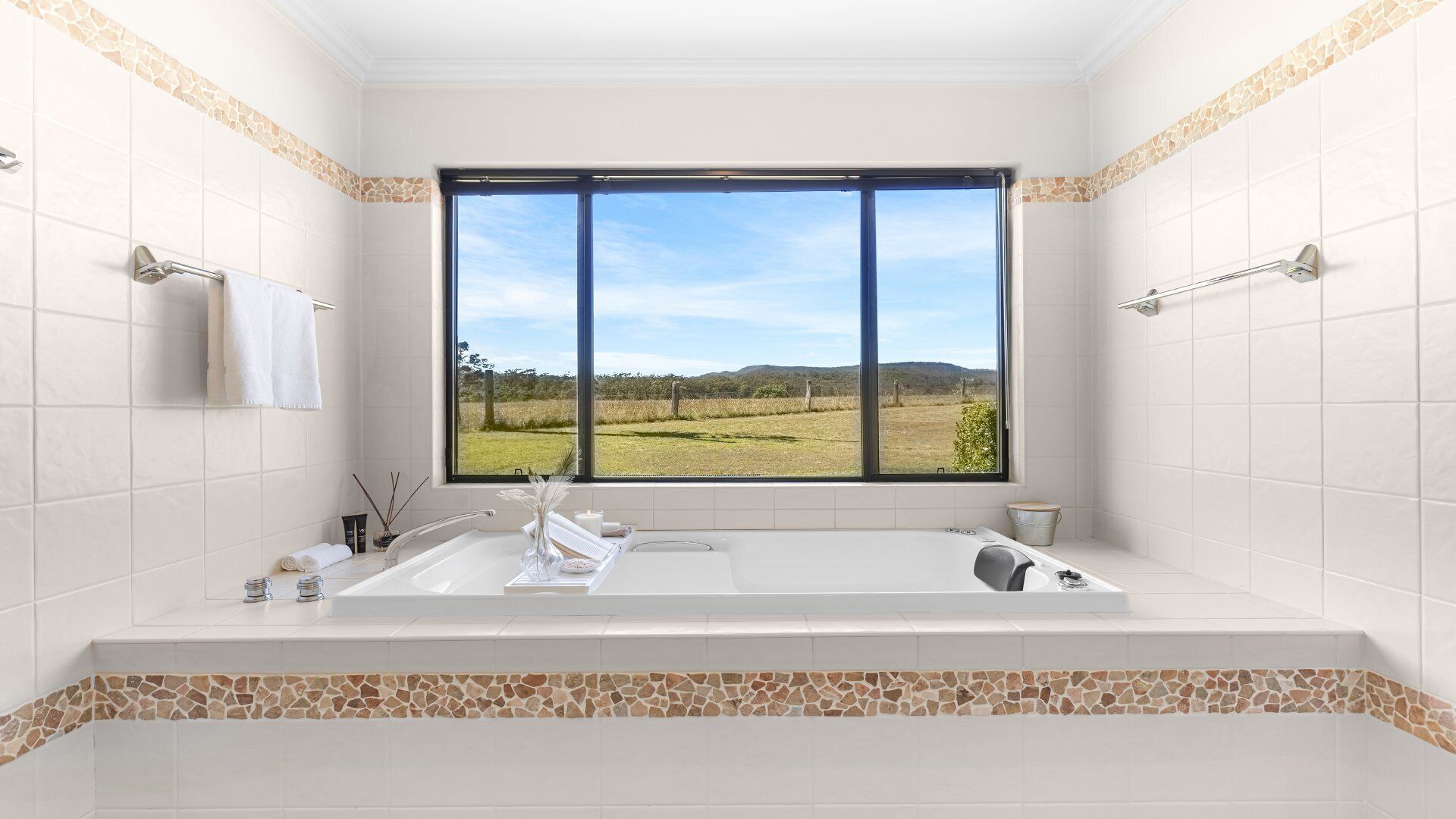 A bathroom with a large bathtub, a window with a view of a field and hills, and tiled walls with a stone border.