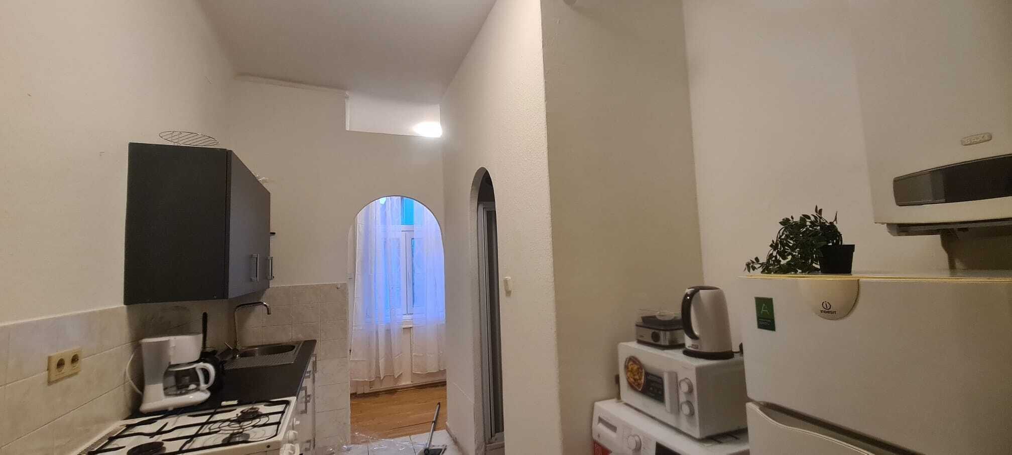 Uma cozinha compacta com fogão a gás, cafeteira, micro-ondas e geladeira. Há uma pequena planta na geladeira e uma janela com cortinas.