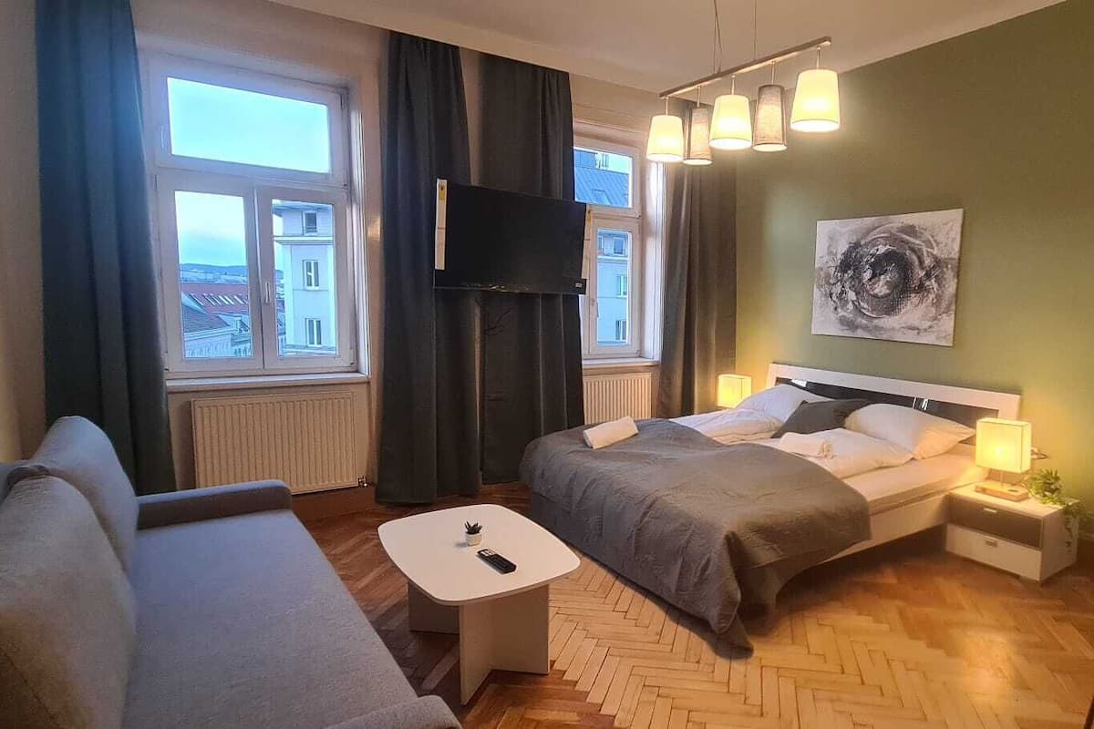 A modern hotel room with a bed, a sofa, a small table, and a desk with chairs.