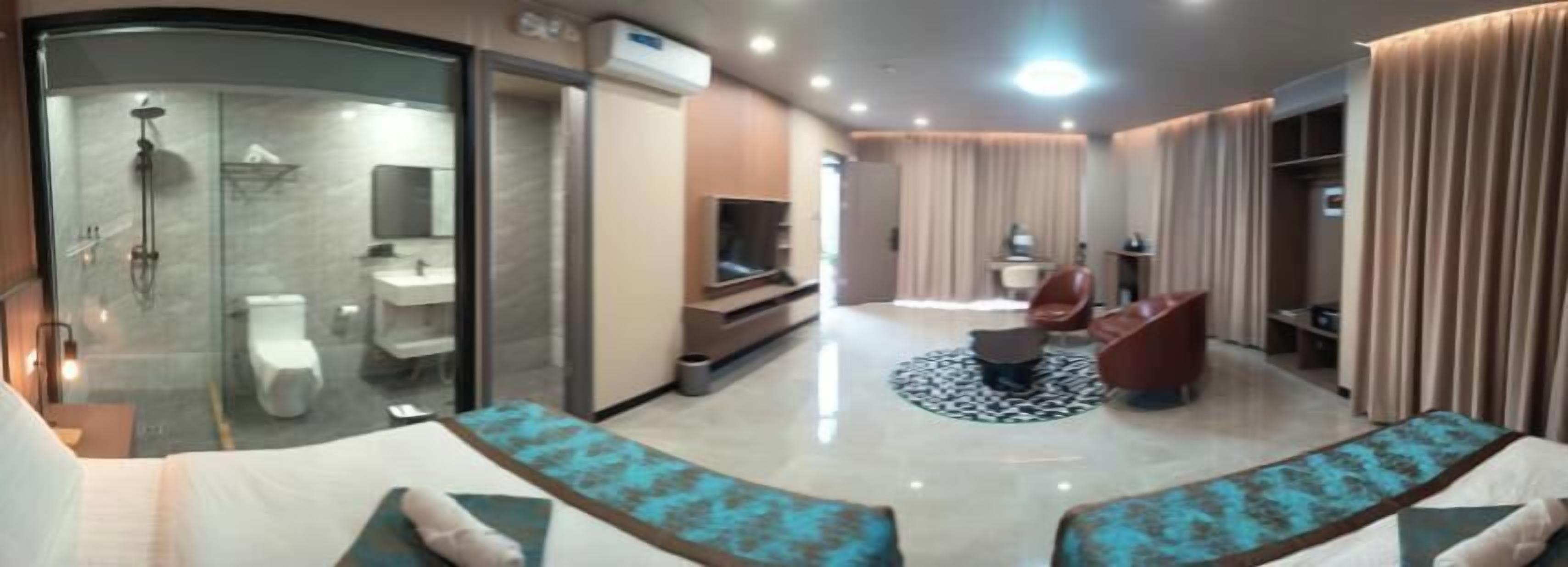 A modern hotel room with a glass shower enclosure, a flat-screen TV, a dining area with a round table, and a seating area with cushioned sofas.