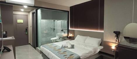 A modern hotel room with a large bed, a glass-enclosed shower, a bedside table, and a built-in mirror.