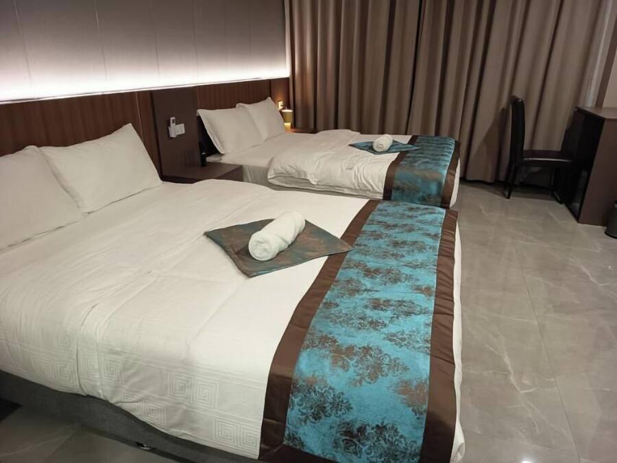 Habitación de hotel con dos camas, cada una con ropa de cama blanca y detalles marrones. Un corredor azul estampado se encuentra entre las camas, y una toalla está sobre él.