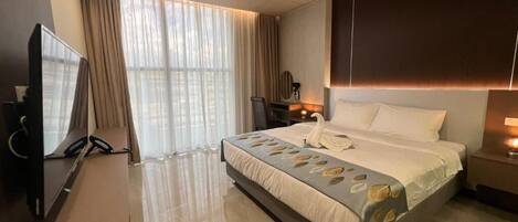 A modern hotel room with a large bed, a flat-screen TV, a desk, and a large window with curtains.