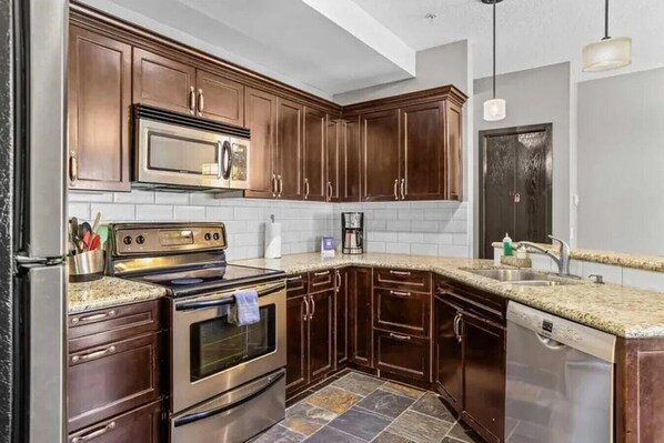 A kitchen with dark wooden cabinets, a stainless steel refrigerator, a double oven, and a granite countertop.