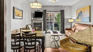 A modern living room with a dining area, a fireplace, a television, and a large window overlooking a garden.