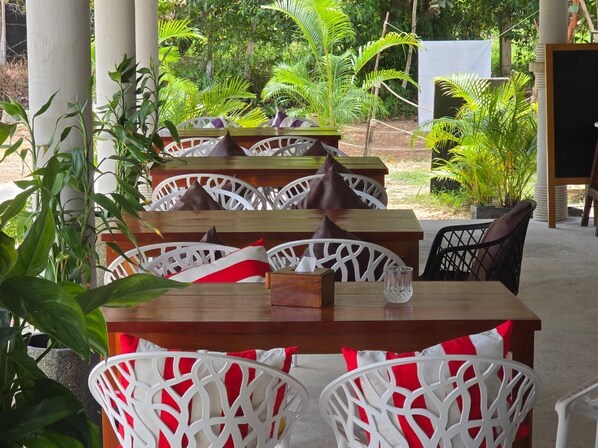 A covered outdoor seating area with wooden tables and white chairs, surrounded by lush greenery. - WHITE BEACH RESORT LONGSET (Koh Rong)