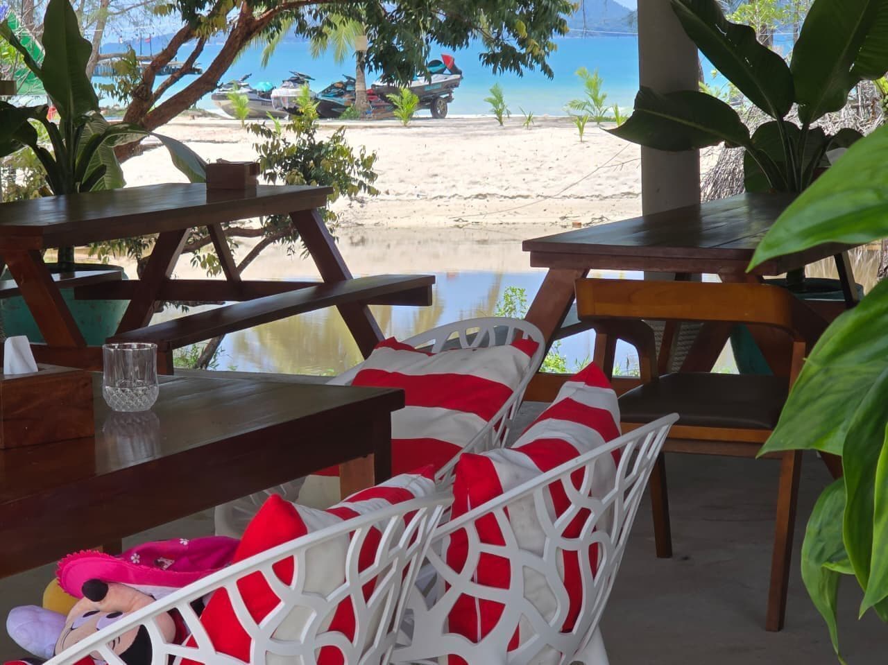 A beachside restaurant with outdoor seating, including white wicker chairs with red cushions, wooden tables, and a view of the beach and ocean.