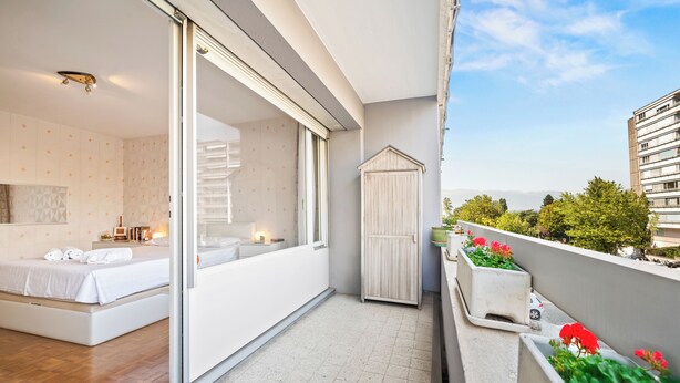 A balcony with a bed, a sliding door, a small wooden cabinet, and potted plants. - Taverney | Spacious, bright apartment (Geneva) (Le Grand-Saconnex)