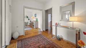 A hallway with a wooden floor, a glass console table with a lamp, a mirror, and a patterned rug. - Taverney | Spacious, bright apartment (Geneva) (Le Grand-Saconnex)