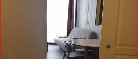 Un pasillo estrecho que conduce a un comedor con un sofá y sillas. Se observa un armario de madera a la izquierda y una puerta a la derecha.