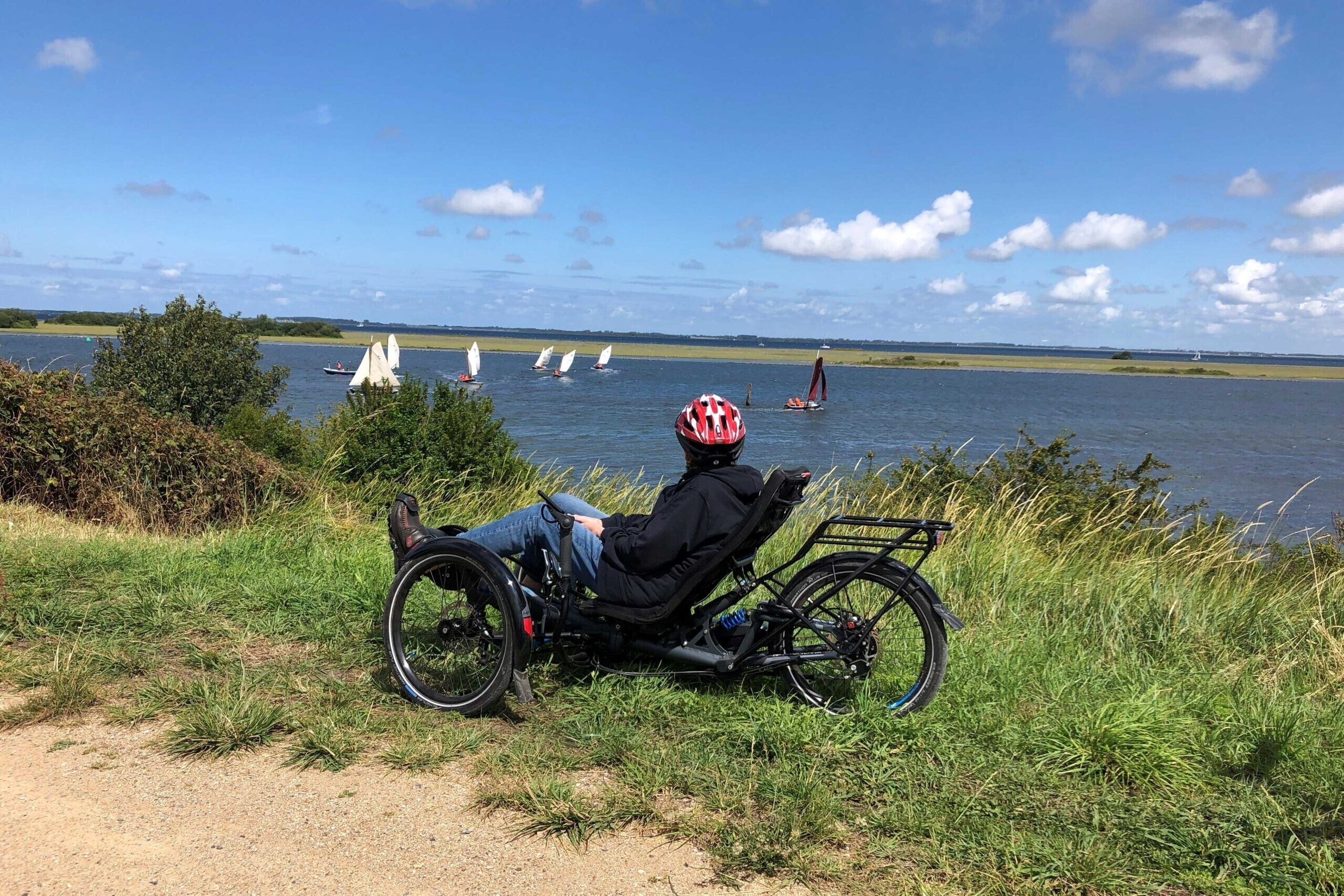 A person sitting on a stationary bike by a lake with sailboats in the background.