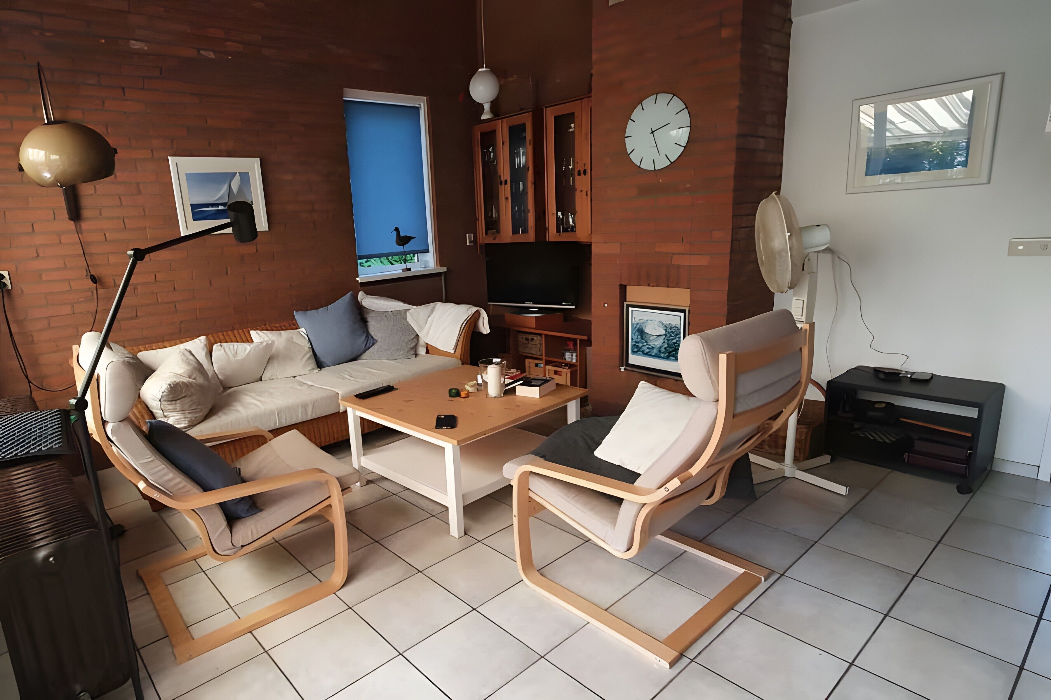 Uma sala de estar com sofá, duas cadeiras de madeira, uma mesa de centro, televisão, relógio e janela com vista.