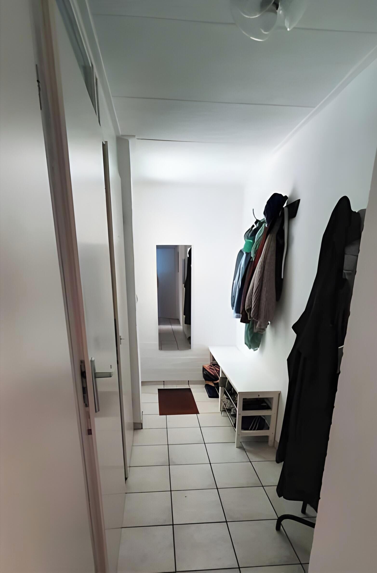 Un pasillo con pared blanca, un perchero con varios objetos, una mesita con espejo y una puerta que da a otra habitación.