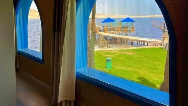 A corridor with arched windows overlooking a waterfront area with a grassy lawn and blue umbrellas. - venecia resort fayoum (Yusuf as-Sidiq)