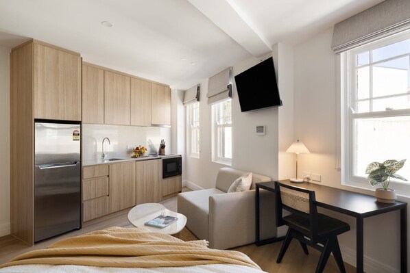 A modern kitchen with a refrigerator, microwave, and wooden cabinets. There is a dining area with a table and chairs, a sofa, and a TV mounted on the wall. - UKO Potts Point (Potts Point)