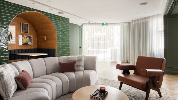 Un moderno vestíbulo de hotel con una pared curva de madera, un sofá, una silla, una mesita y un gran ventanal con cortinas.