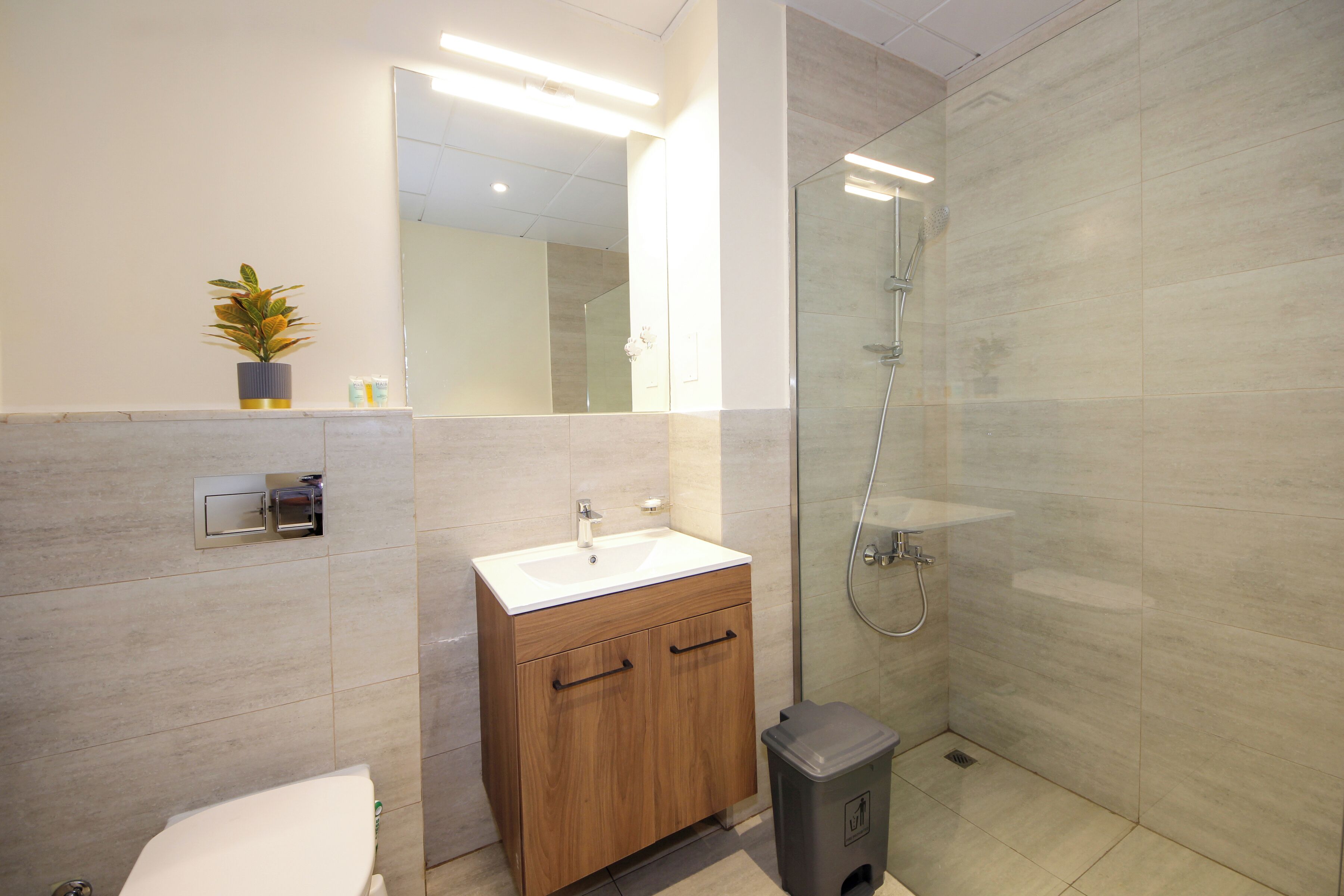 A modern bathroom with a wooden vanity, a glass shower enclosure, and a small trash bin.