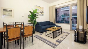 A modern living room with a blue sofa, wooden dining table, and chairs, a coffee table, a large potted plant, and a view of a building through a glass door. - Silk Valley-Luxury 1B near Masdar Park (Abu Dhabi)