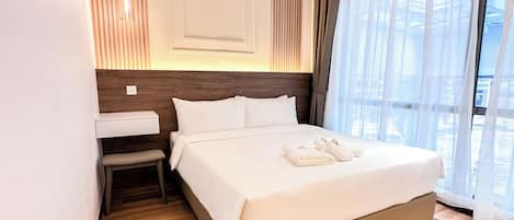 A hotel room with a bed, a bedside table, a chair, and a window with sheer curtains.
