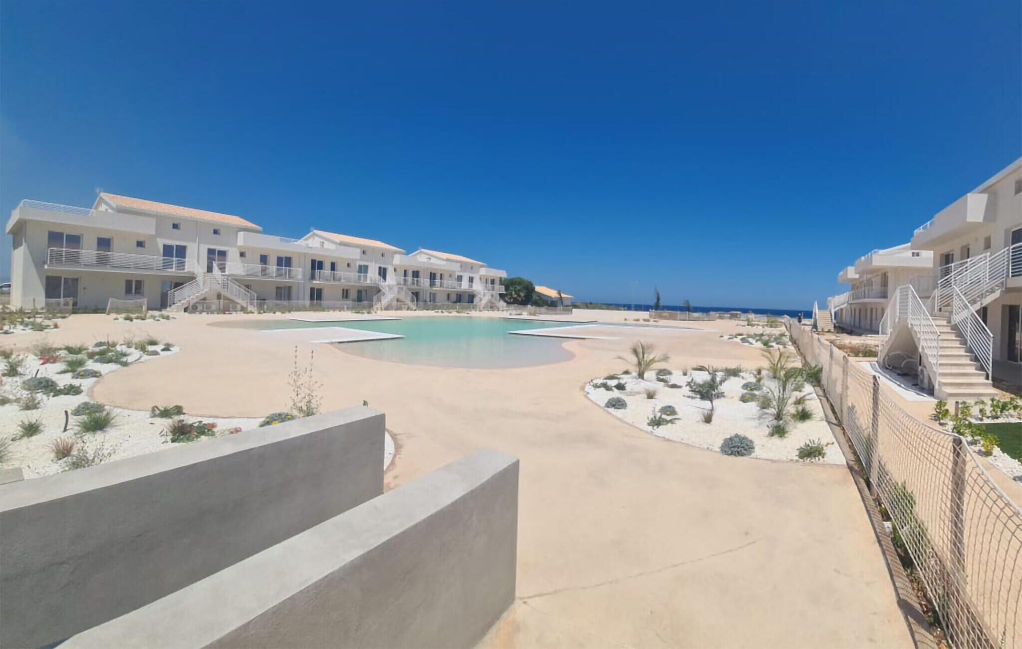 A resort with a central pool, multiple buildings with balconies, and a clear view of the sea.