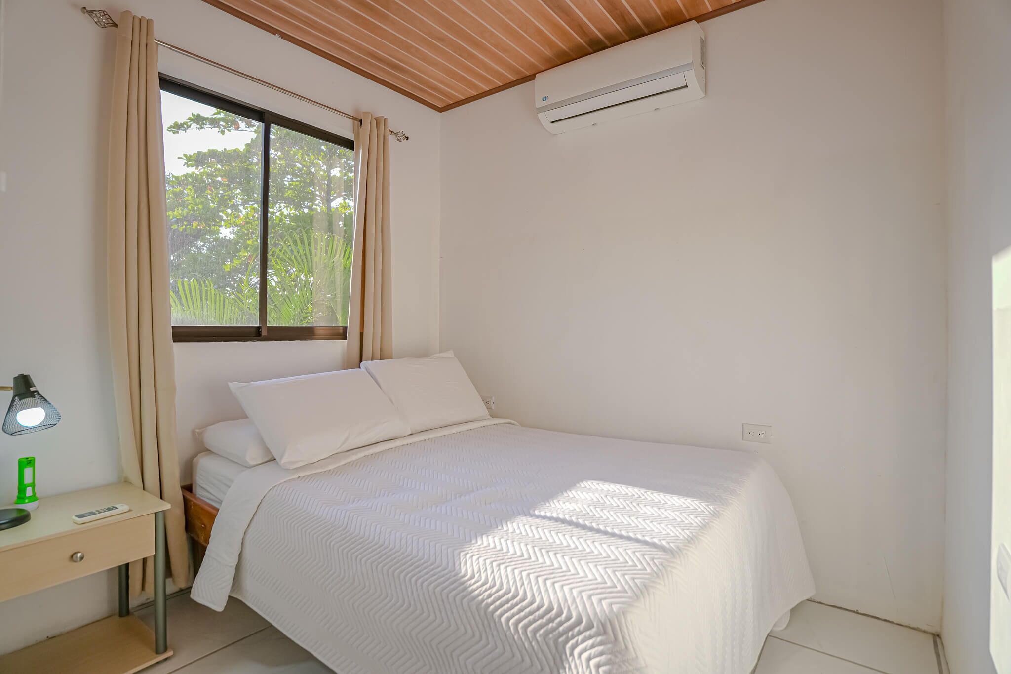 A bedroom with a bed, a desk, a lamp, a window with curtains, and an air conditioning unit.