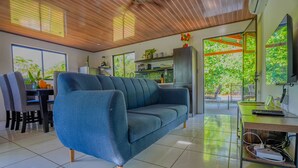 Un salon moderne avec un canapé bleu, un coin repas avec des chaises, une cuisine équipée d’un réfrigérateur et une grande fenêtre donnant sur un espace verdoyant.