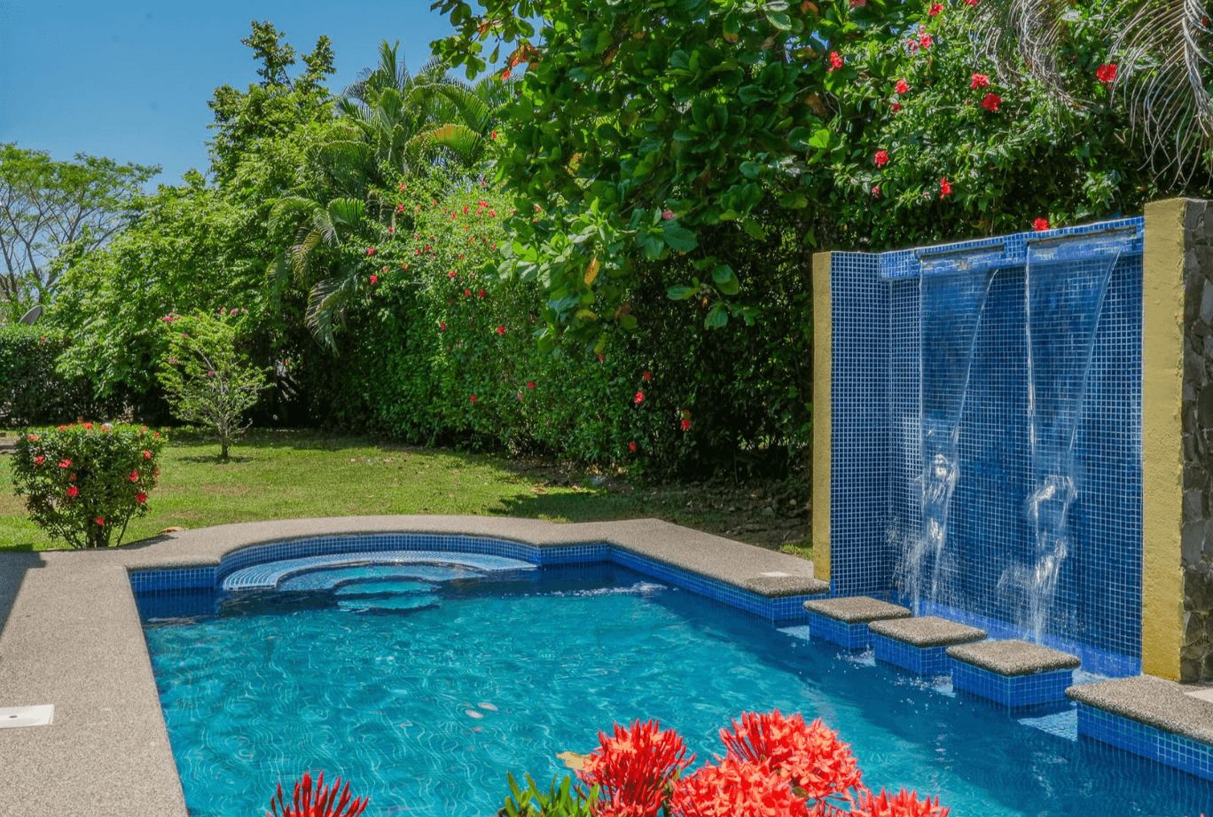 Une piscine dont le bord est recouvert de carreaux bleus, entourée d’une végétation luxuriante et de fleurs rouges.