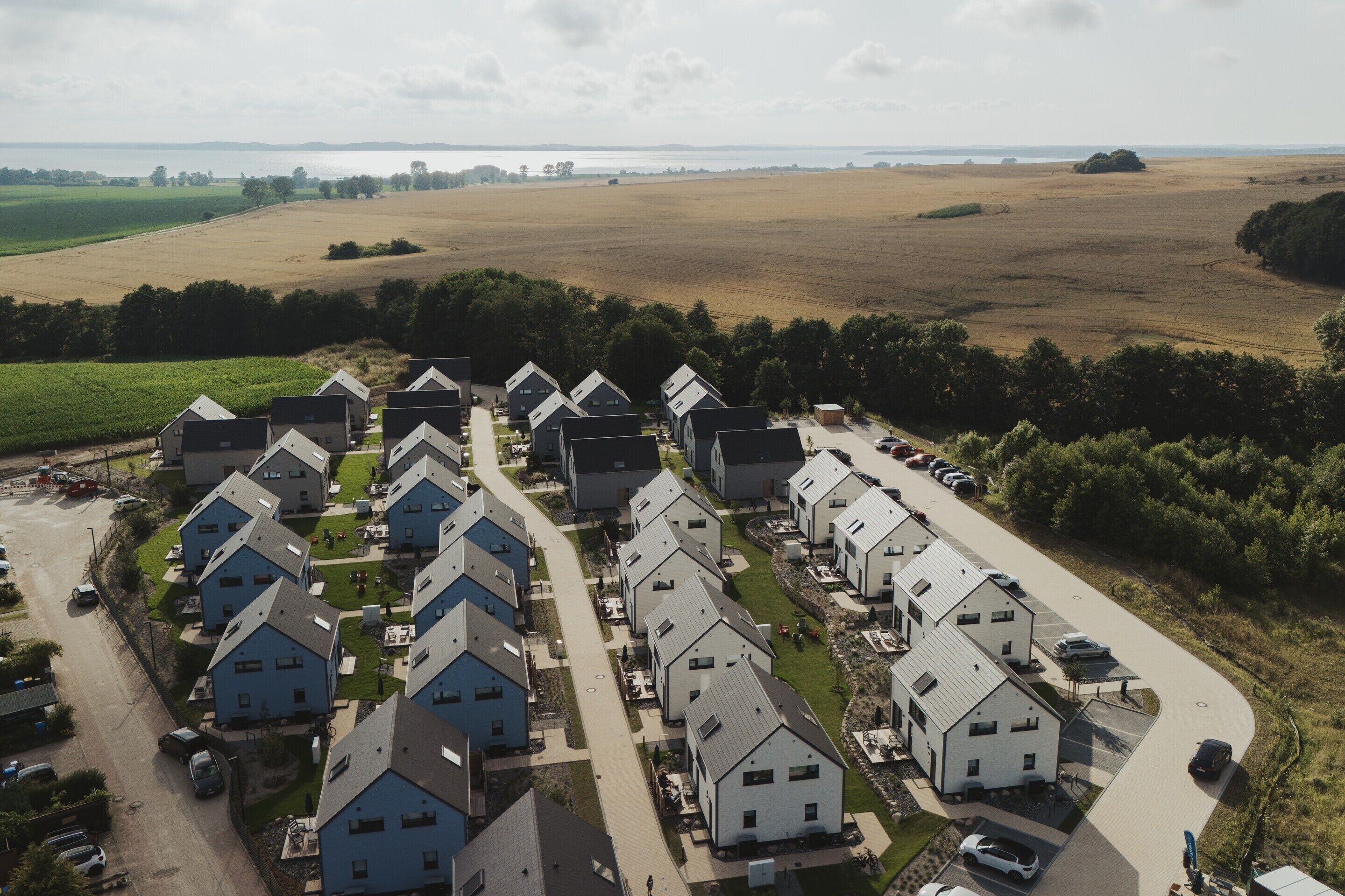 A residential area with multiple houses arranged in rows, a parking area, and a view of a large open field and water body in the background.