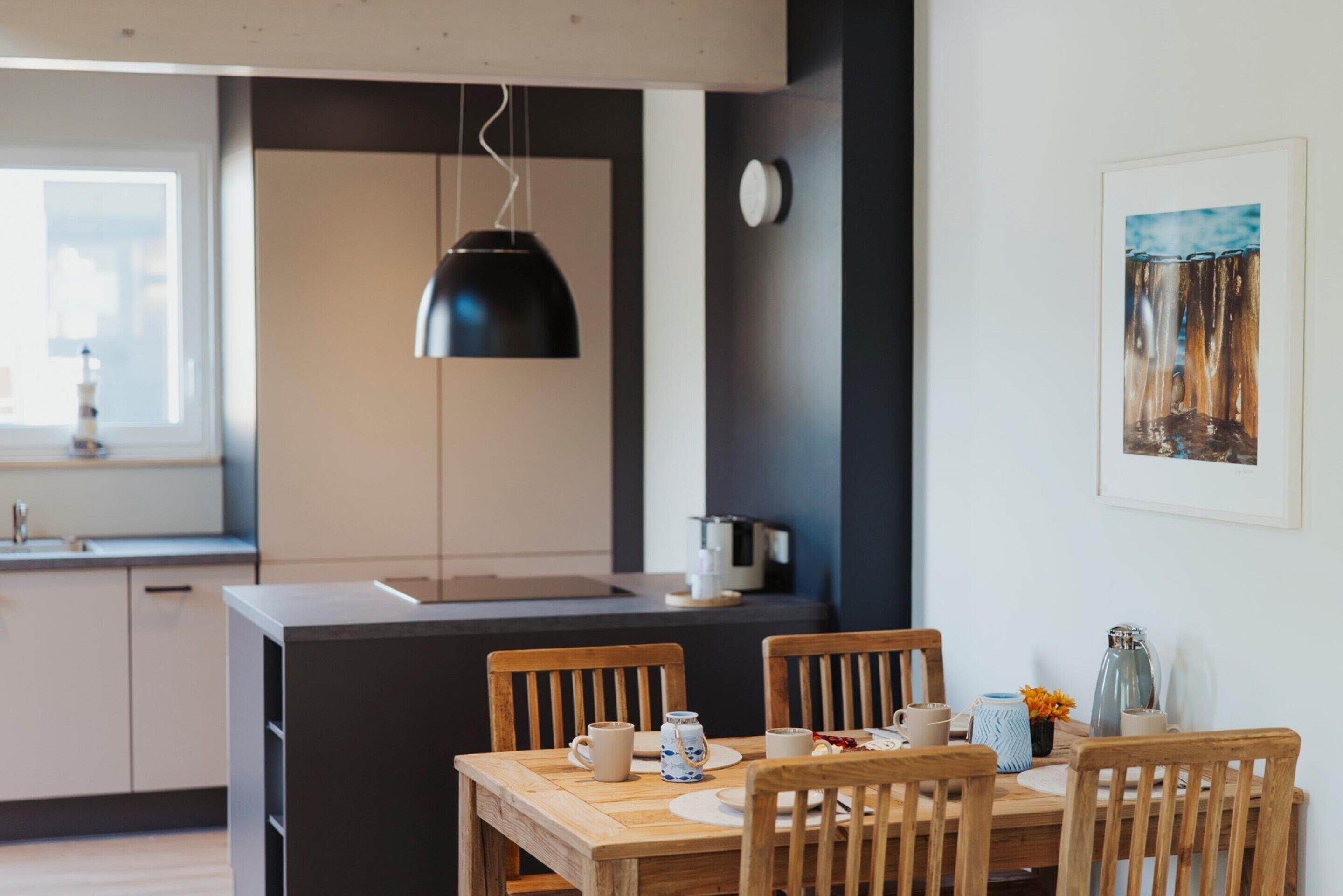 Une cuisine moderne avec des meubles en bois, un luminaire suspendu noir et un coin repas avec des chaises et une table en bois, le tout disposé pour deux personnes.