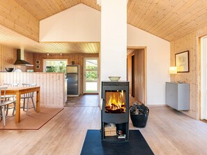 A modern cabin interior with a central fireplace, wooden walls, a kitchen area, and a dining space.