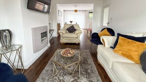Un salon moderne avec un canapé, un fauteuil et une table basse. On y trouve une cheminée, un téléviseur fixé au mur et, au fond, un coin repas.