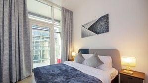 A hotel room with a large bed, a nightstand with a lamp, a window with curtains, and a wall-mounted abstract art piece.