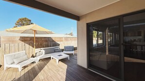 En overdækket udendørs terrasse med træterrasse, hvide møbler og stor parasol.