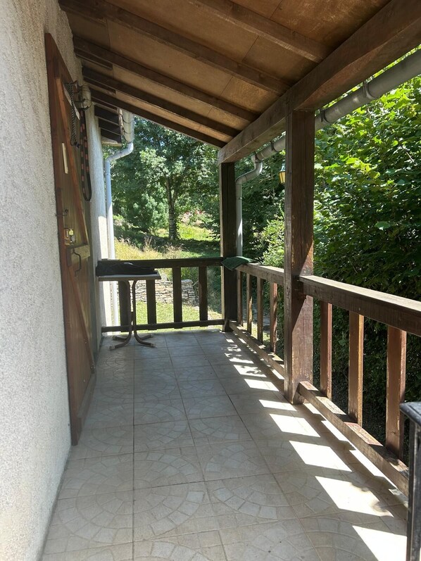Un espacio al aire libre techado, con suelo de baldosas, baranda de madera y vistas a la vegetación.