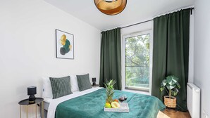 A neatly arranged bedroom with a bed, a bedside table with a lamp, a window with curtains, a framed picture on the wall, and a tray of fruit and wine on the bed.
