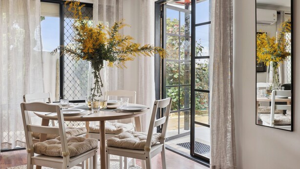Un coin repas avec une table ronde, des chaises blanches et un vase rempli de fleurs jaunes.