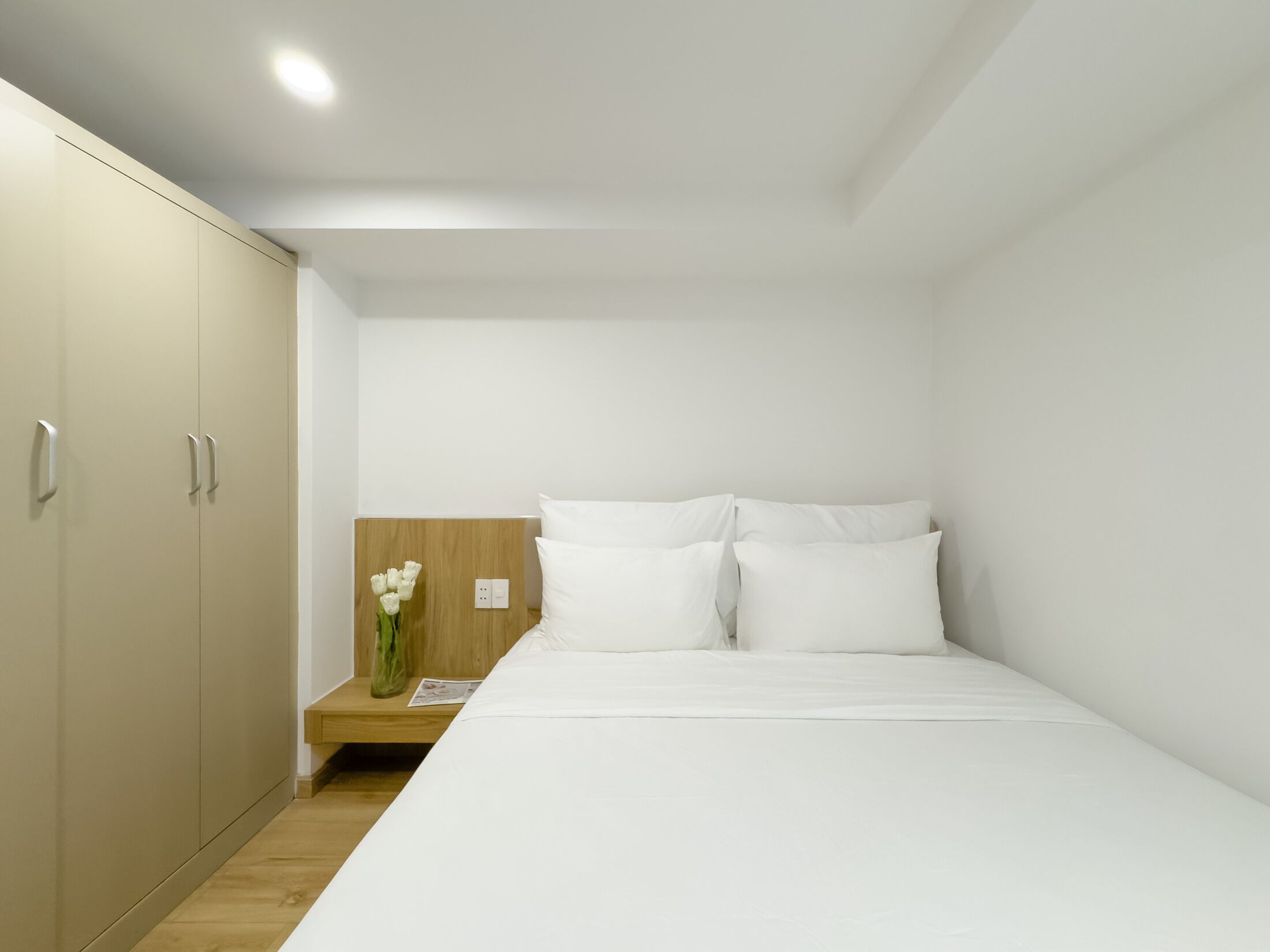 A minimalist hotel room with a bed, a wooden nightstand, and a wardrobe.