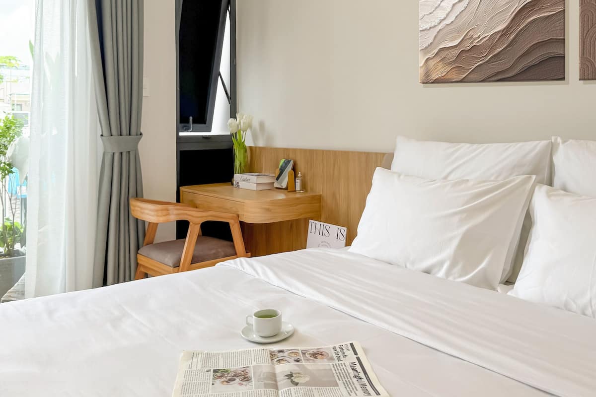 Un lit bien fait, avec du linge de lit blanc, une table de chevet sur laquelle se trouvent une tasse de café, un journal et une pancarte sur laquelle on peut lire : « ICI ».