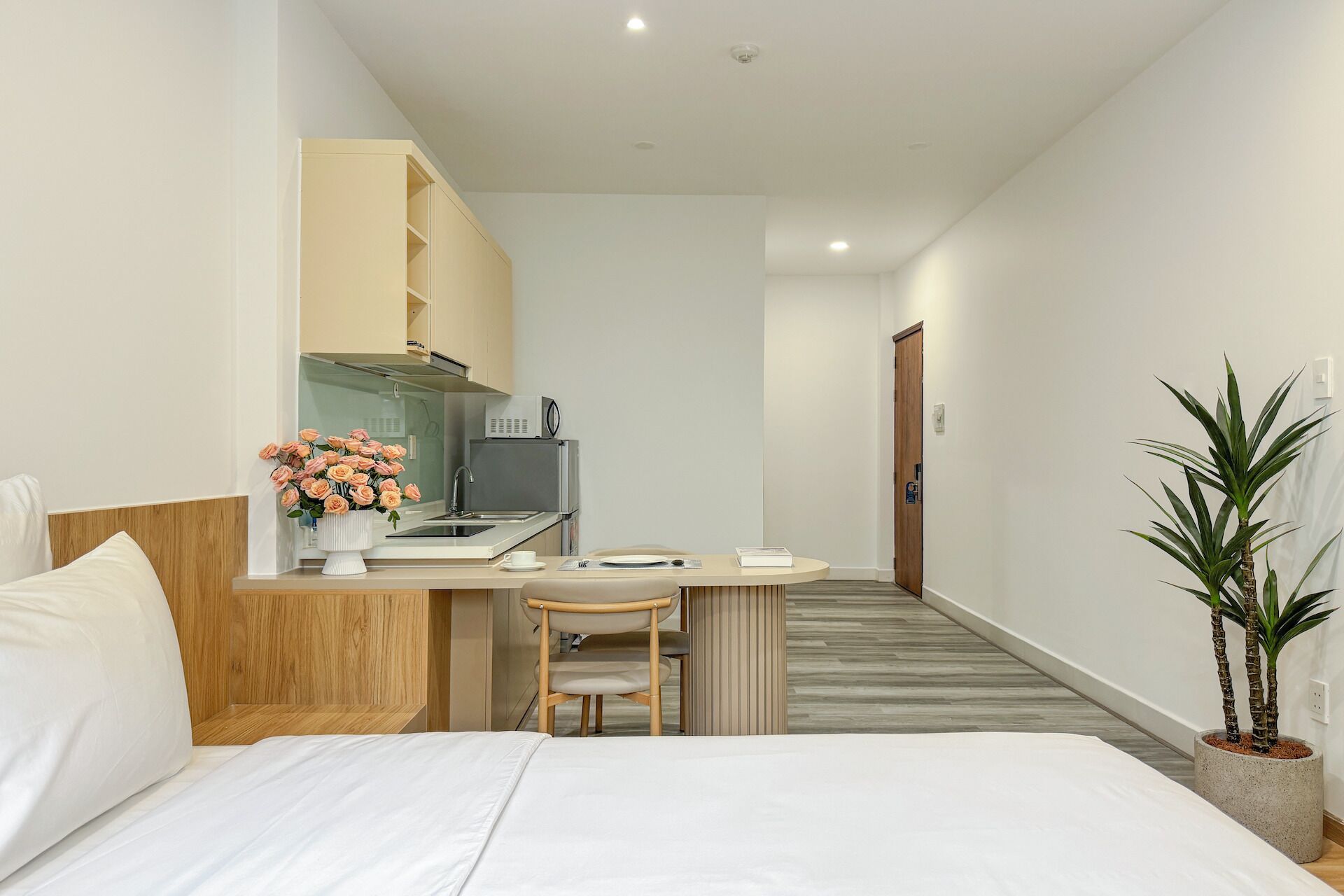 A hotel room with a bed, a desk with a chair, a small kitchenette, and a potted plant.
