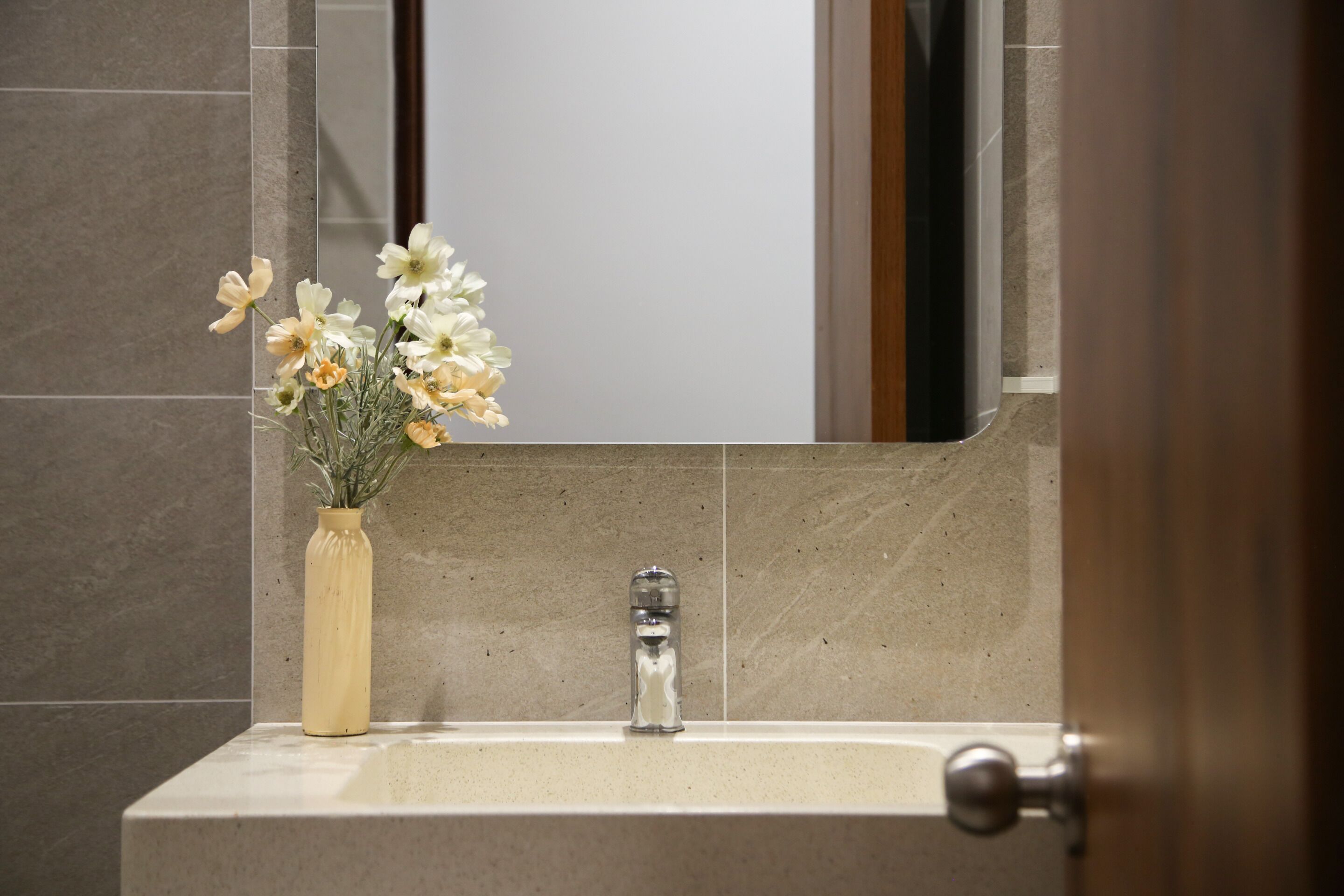 A bathroom with a beige sink, a mirror above it, and a vase with flowers on the left.