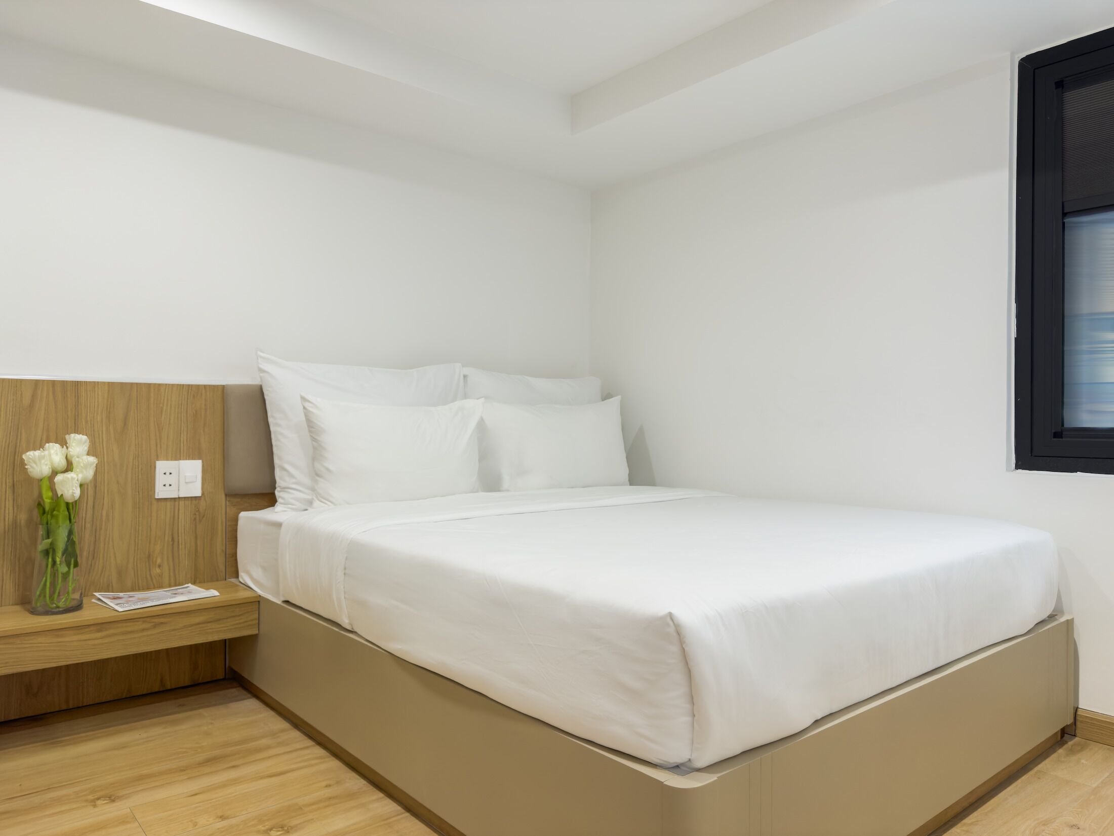 A minimalist bedroom with a bed, a nightstand, a vase with flowers, and a window with blinds.