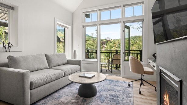 A modern living room with a grey sofa, a round coffee table, and a fireplace. Large windows offer a view of greenery and a balcony. - Skyline Loft | Capitol Peak Lodge #3323 | 1B/1B (Snowmass Village)
