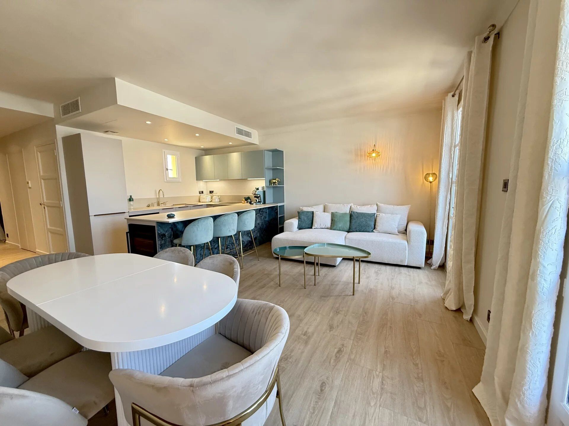 A modern living room with a white sofa, a dining area with a white table and chairs, and a kitchen with grey cabinets and a central island.