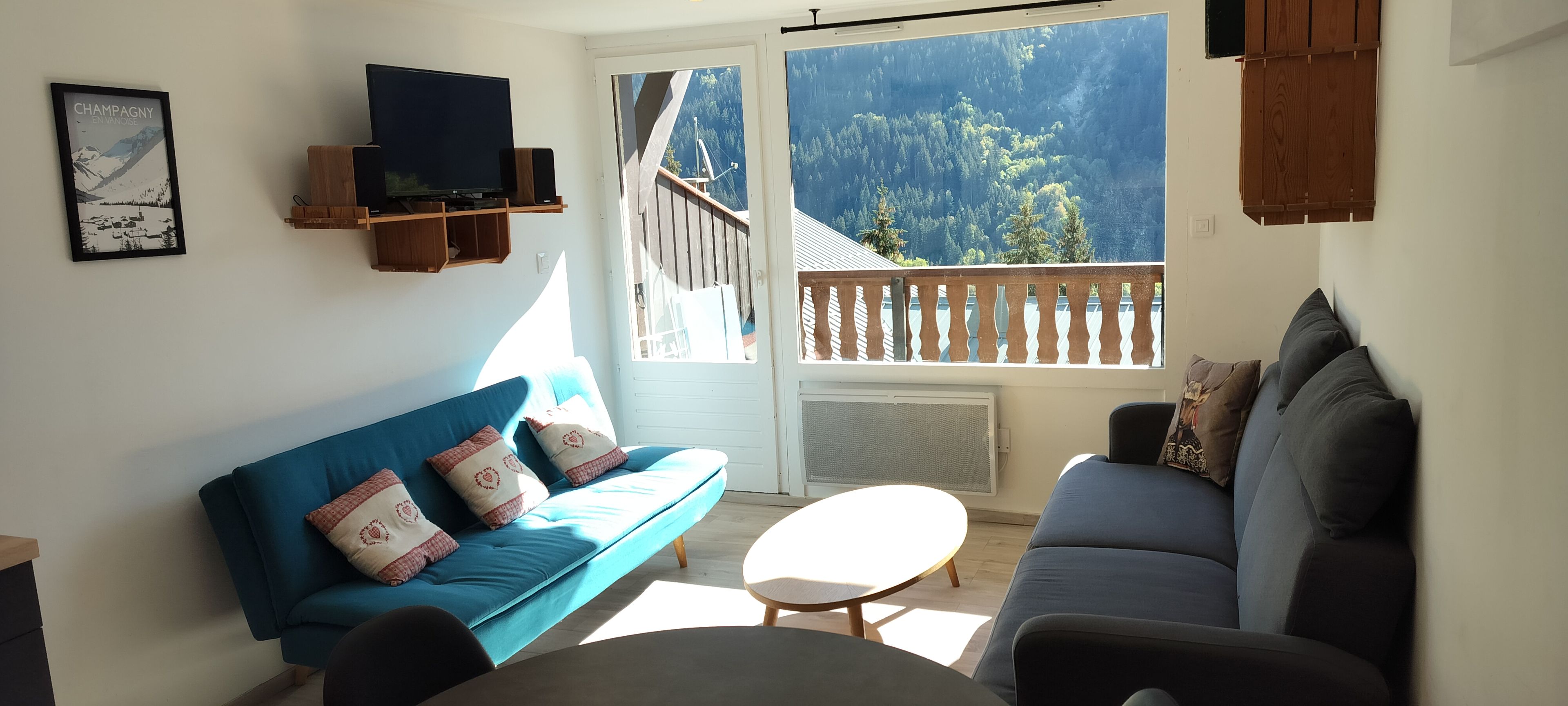 A living room with a blue sofa, a grey sofa, and a small table. There is a TV mounted on the wall and a window with a view of trees.