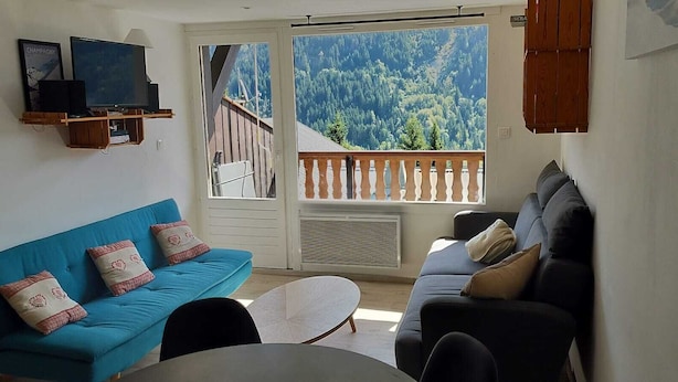 A living room with a blue sofa, a dark sofa, and a white coffee table. There is a TV on a wooden shelf and a large window with a view of a forested mountain. - 6-8 person apartment at the foot of the lifts, in a typical village (Champagny-en-Vanoise)