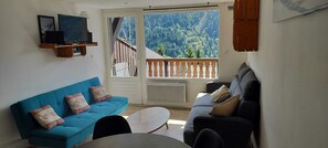 Una sala de estar con un sofá azul, un sofá oscuro y una mesa de centro blanca. Hay un televisor en un estante de madera y un ventanal con vistas a una montaña boscosa.