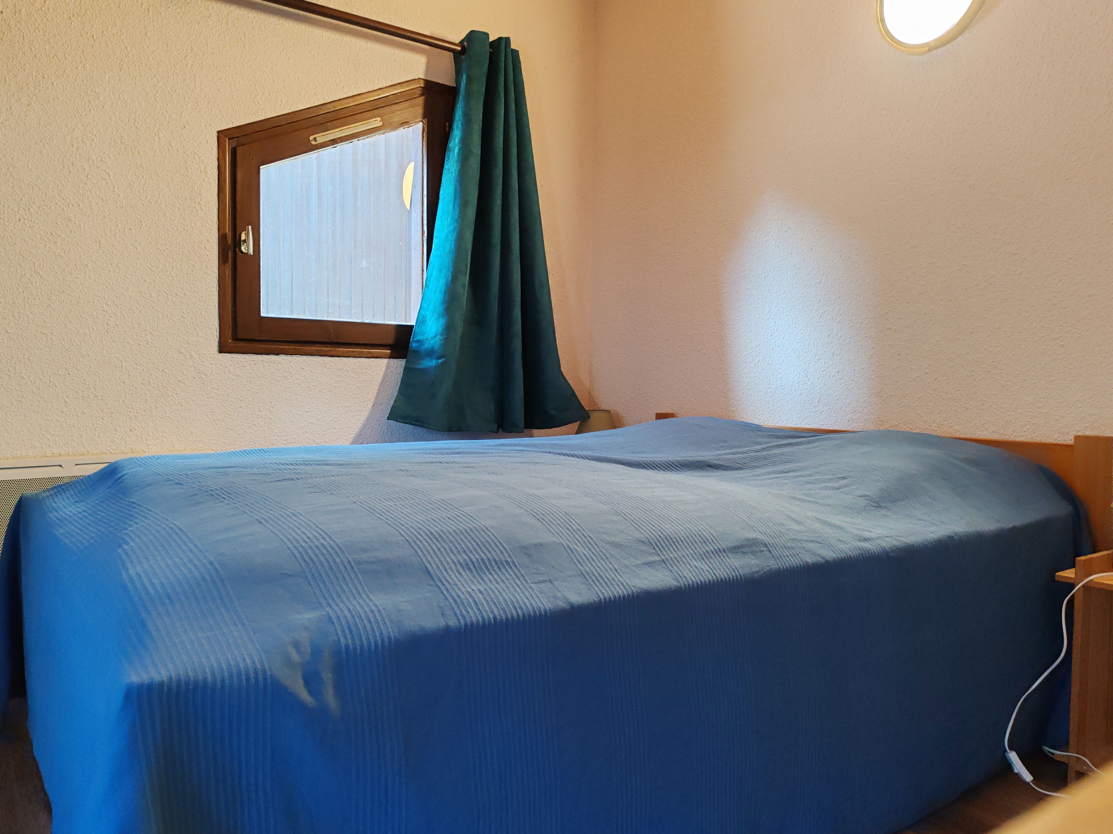 Un llit individual amb un edredó blau, una finestra amb cortina i una làmpada de paret.