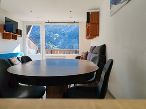 Un coin repas avec une table ronde et des chaises, un balcon avec vue sur les montagnes et un téléviseur fixé au mur.