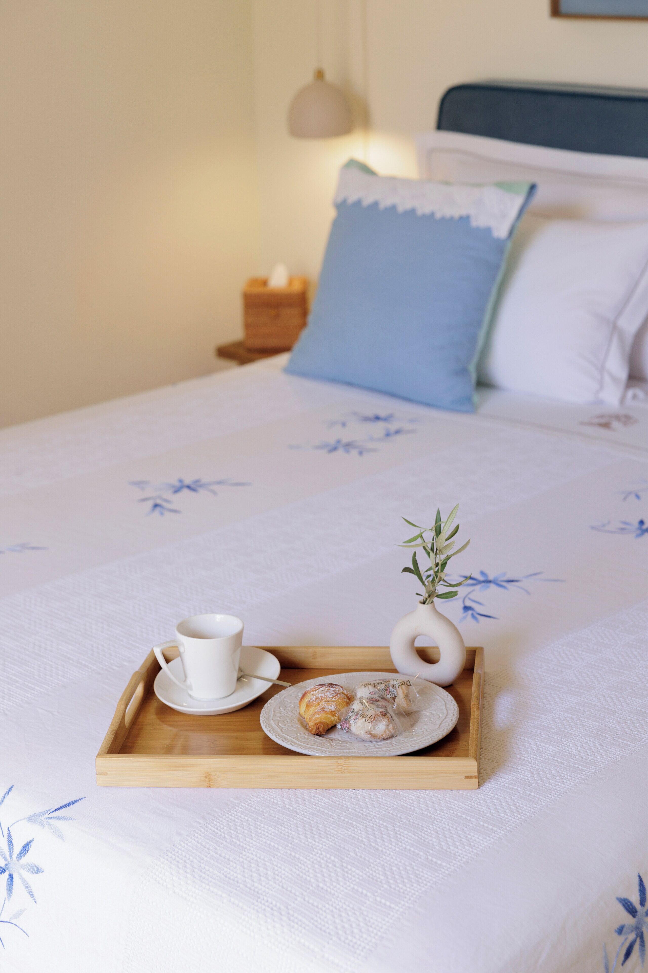 Un lit bien fait, sur lequel est posé un plateau contenant divers éléments pour le petit-déjeuner, notamment une tasse, une assiette de viennoiseries et un petit vase avec une plante.