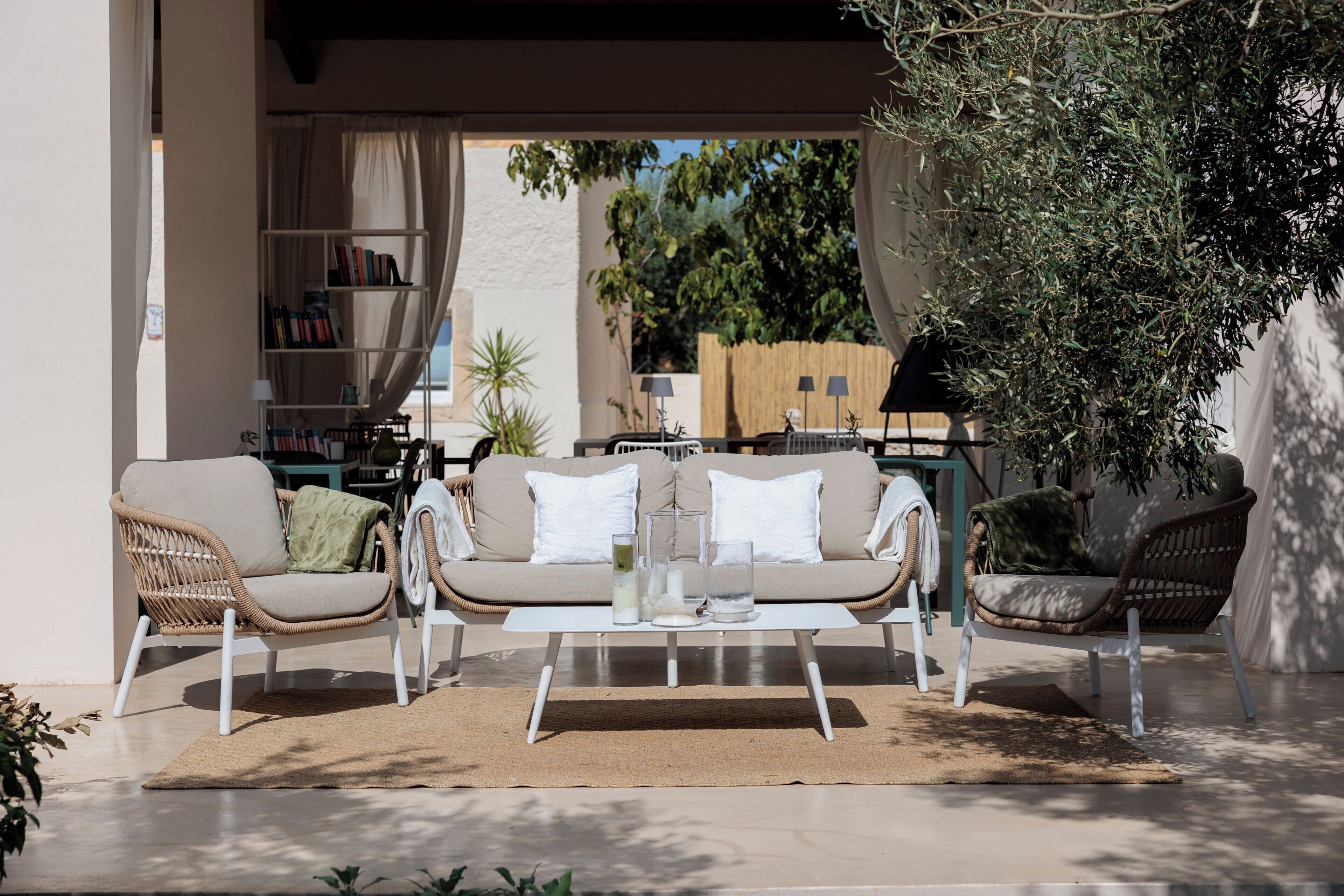 A sunlit outdoor seating area with a sofa, chairs, and a coffee table, surrounded by bookshelves and greenery.